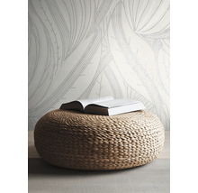 Vliesbehang met groot bladmotief in een lichte, moderne kamer met een gevlochten poef van natuurlijk materiaal.