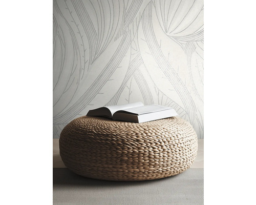 Vliesbehang met groot bladmotief in een lichte, moderne kamer met een gevlochten poef van natuurlijk materiaal.