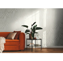 Licht vliesbehang met grafisch bladpatroon in een modern ingerichte woonkamer met een oranje bank, bijzettafel en kamerplant.