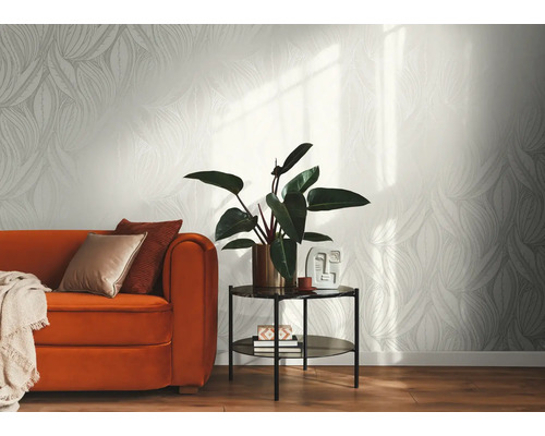 Licht vliesbehang met grafisch bladpatroon in een modern ingerichte woonkamer met een oranje bank, bijzettafel en kamerplant.