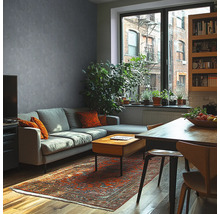 Moderne woonkamer met grijze muur in betonlook, grijze bank, houten tafel, oosters tapijt en grote kamerplanten voor een raam.