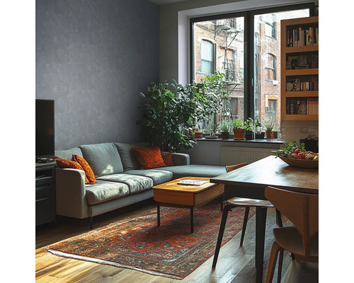 Moderne woonkamer met grijze muur in betonlook, grijze bank, houten tafel, oosters tapijt en grote kamerplanten voor een raam.