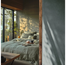 Moderne slaapkamer met groen linnen beddengoed, grote glazen pui met uitzicht op planten en een muur met betonlook en schaduweffecten.