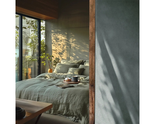 Moderne slaapkamer met groen linnen beddengoed, grote glazen pui met uitzicht op planten en een muur met betonlook en schaduweffecten.