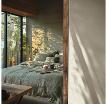 Moderne slaapkamer met bed in groen linnen beddengoed, muur in betonlook met boomschaduwen en groot raam met planten.