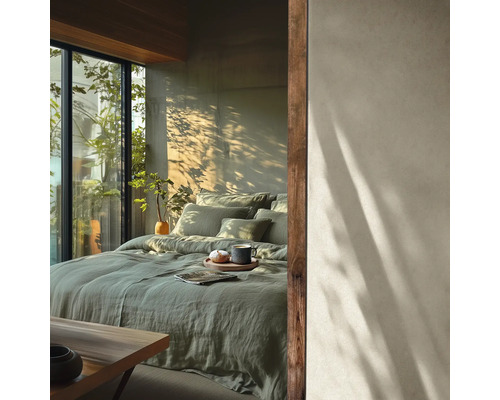 Moderne slaapkamer met bed in groen linnen beddengoed, muur in betonlook met boomschaduwen en groot raam met planten.