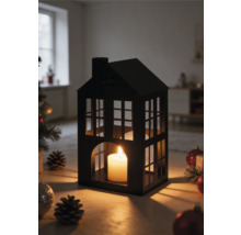 Lantaarn in de vorm van een huis van zwart metaal met een brandende kaars, omringd door kerstversiering en dennenappels in een lichte kamer.