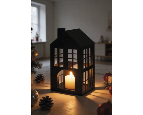 Lantaarn in de vorm van een huis van zwart metaal met een brandende kaars, omringd door kerstversiering en dennenappels in een lichte kamer.