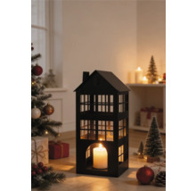 Zwarte metalen lantaarn in de vorm van een huis met een brandende kaars in een kerstscène met een versierde boom en cadeaus.