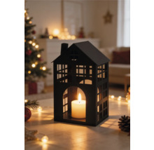 Zwarte metalen lantaarn in de vorm van een huis met brandende kaars in een lichte woonkamer met kerstboom en lichtjes.
