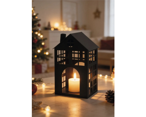 Zwarte metalen lantaarn in de vorm van een huis met brandende kaars in een lichte woonkamer met kerstboom en lichtjes.