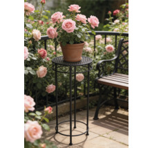 Hoge ronde plantenstandaard van zwart metaal met ornamenten met daarop een terracotta pot met roze rozen in een zonnige tuin naast een bank.