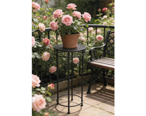 Hoge ronde plantenstandaard van zwart metaal met ornamenten met daarop een terracotta pot met roze rozen in een zonnige tuin naast een bank.