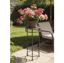 Hoge decoratieve metalen plantenstandaard op een zonnig tuinterras met een ronde geperforeerde bovenkant en krulornamenten.