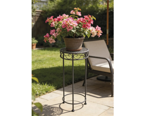 Hoge decoratieve metalen plantenstandaard op een zonnig tuinterras met een ronde geperforeerde bovenkant en krulornamenten.