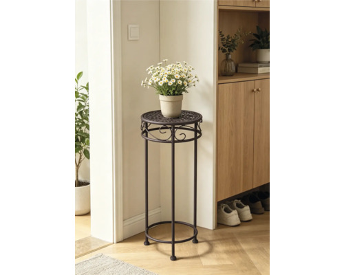 Hoge plantenstandaard van donker metaal met decoratieve ornamenten en witte bloemen in een lichte kamer naast een houten kast.