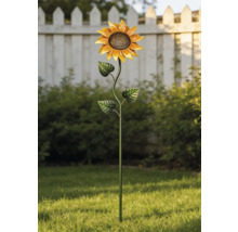 Metalen tuinsteker in de vorm van een zonnebloem in een lichte tuin op een groen gazon voor struiken en een wit hek.