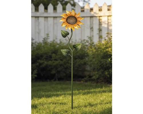 Metalen tuinsteker in de vorm van een zonnebloem in een lichte tuin op een groen gazon voor struiken en een wit hek.
