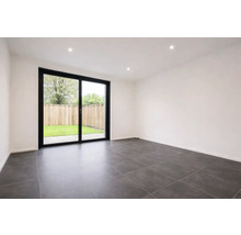 Moderne, lichte kamer met donkergrijze vloertegels, witte muren en een grote glazen schuifdeur met zwart frame naar de tuin.
