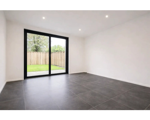 Moderne, lichte kamer met donkergrijze vloertegels, witte muren en een grote glazen schuifdeur met zwart frame naar de tuin.