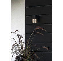 Moderne zwarte buitenwandlamp met verticale lichtgleuven aan een donkere houten wand naast siergras.