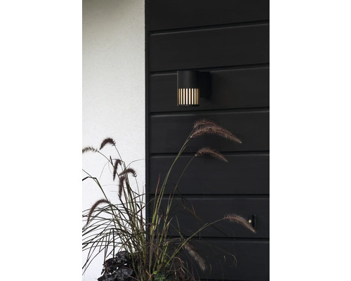 Moderne zwarte buitenwandlamp met verticale lichtgleuven aan een donkere houten wand naast siergras.