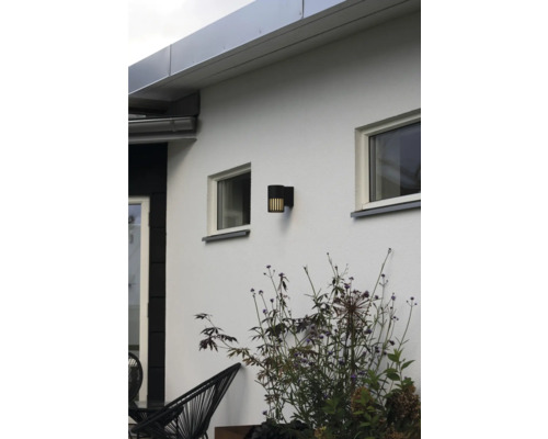 Moderne zwarte cilindrische buitenwandlamp met verticale lichtlamellen op een witte buitenmuur tussen ramen en planten.