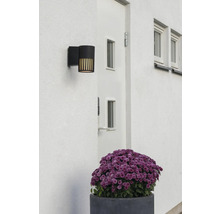 Moderne zwarte buitenwandlamp op een witte gevel naast een deur, voorzien van verticale lichtsleuven.
