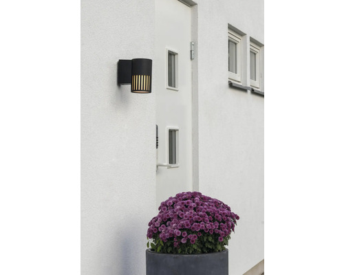 Moderne zwarte buitenwandlamp op een witte gevel naast een deur, voorzien van verticale lichtsleuven.