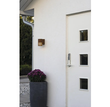 Moderne entree met witte voordeur, digitaal deurslot, buitenwandlamp met roest-look en een grote plantenbak met paarse bloemen.