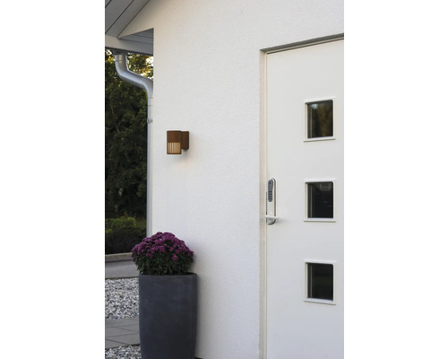 Moderne entree met witte voordeur, digitaal deurslot, buitenwandlamp met roest-look en een grote plantenbak met paarse bloemen.