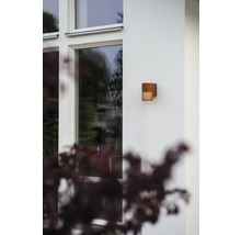 Moderne buitenwandlamp van metaal in roestlook op een lichte huismuur naast een raam.