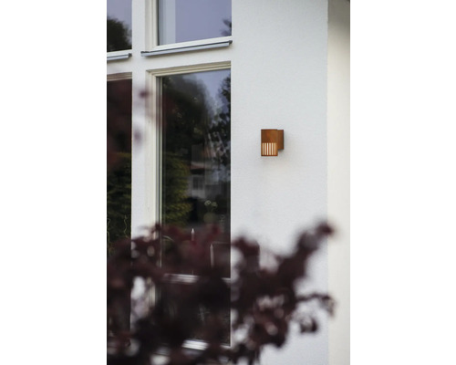 Moderne buitenwandlamp van metaal in roestlook op een lichte huismuur naast een raam.