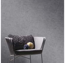 Grijs vliesbehang met betonlook op een muur achter een moderne fauteuil met structuurkussen en decoratieve papieren bollen.