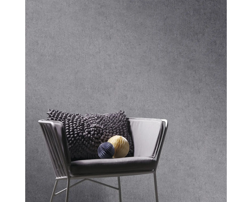 Grijs vliesbehang met betonlook op een muur achter een moderne fauteuil met structuurkussen en decoratieve papieren bollen.