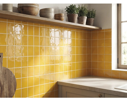 Keukenachterwand met gele glanzende vierkante wandtegels, een houten plank met schalen en kruiden en een aanrechtblad.