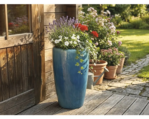 Hoge blauwe keramieken plantenbak met lavendel en geraniums op een houten terras naast een houten tuinhuis.