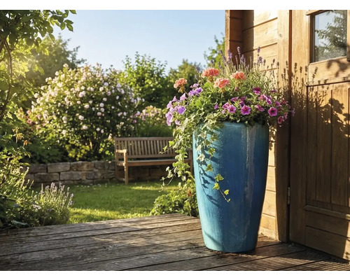 Hoge blauwe keramische plantenbak met zomerbloemen en klimop op een houten terras in een lichte tuin met tuinbank en stenen muur.
