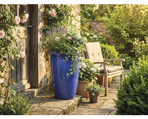 Hoge blauwe keramische bloempot op stenen terras naast rustieke houten deur, tuinstoel en bloemen in een lichte, verzorgde tuin.