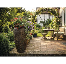 Beplante hoge plantenbak op een geplaveid terras met houten tuinmeubelen, rozenboog en vakwerkhuis op de achtergrond.