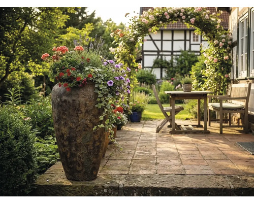 Beplante hoge plantenbak op een geplaveid terras met houten tuinmeubelen, rozenboog en vakwerkhuis op de achtergrond.