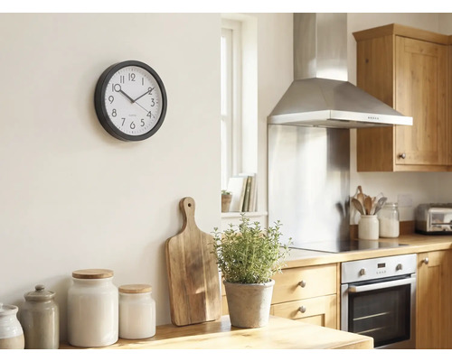 Lichte keuken met een ronde wandklok aan de muur, houten snijplank, kruidenpot en voorraadpotten op het aanrecht.