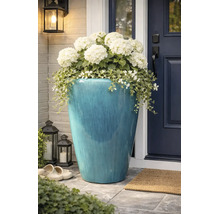 Grote, blauw geglazuurde keramische plantenpot met witte hortensia's en hangplanten bij een donkere voordeur.