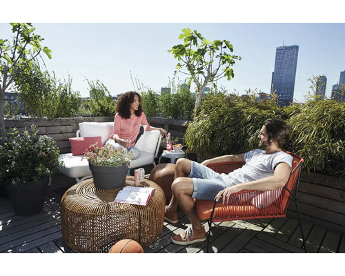 Licht dakterras met loungemeubels, waaronder een ronde vlechttafel, een witte buitenbank, een rode stoel en diverse groene planten zoals bamboe.