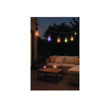 Gekleurde LED-lichtsnoer boven een loungeset met bank, kussens en kaarsen op een houten tafel in een tuin bij nacht.