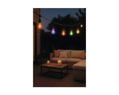 Gekleurde LED-lichtsnoer boven een loungeset met bank, kussens en kaarsen op een houten tafel in een tuin bij nacht.