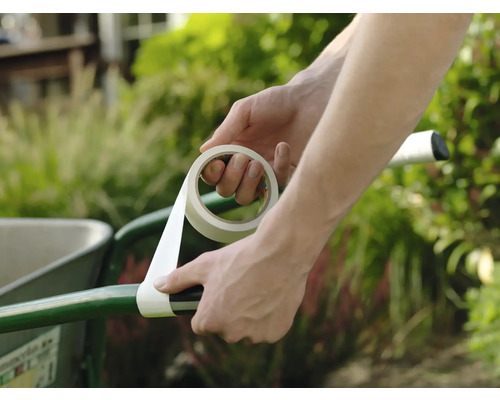 Iemand wikkelt witte tape om de handgreep van een groene kruiwagen in de tuin ter reparatie.