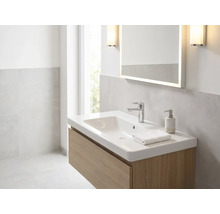 Lichte badkamer met witte wastafel op houten onderkast, verchroomde kraan, verlichte spiegel en wandlampen.