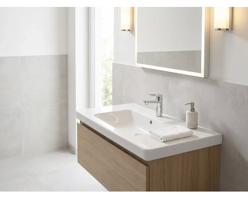 Lichte badkamer met witte wastafel op houten onderkast, verchroomde kraan, verlichte spiegel en wandlampen.