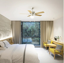 Lichte, moderne slaapkamer met plafondventilator met licht, vijf houten bladen, bed met wit beddengoed en twee gele fauteuils.
